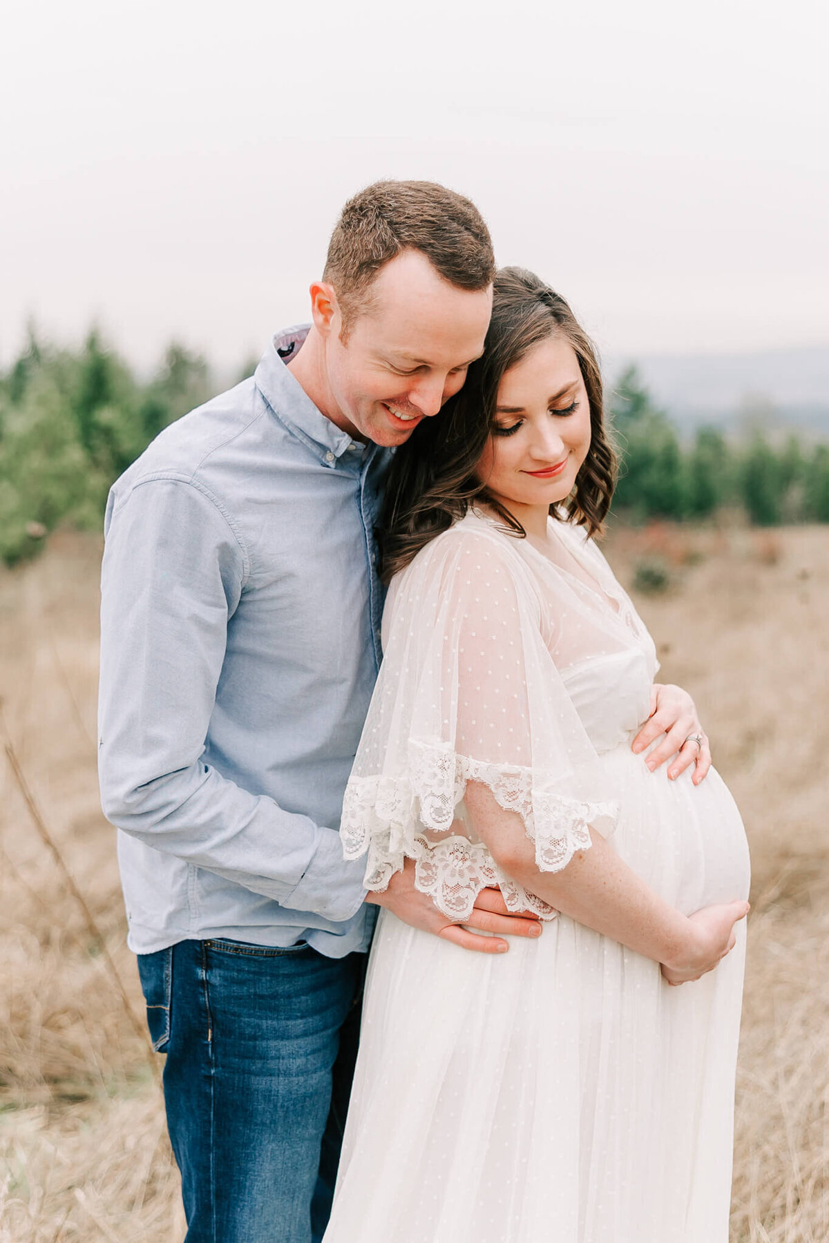mom wearing white dress happily holding her pregnant belly with her husband behind her who is wearing a blue shirt and jeans