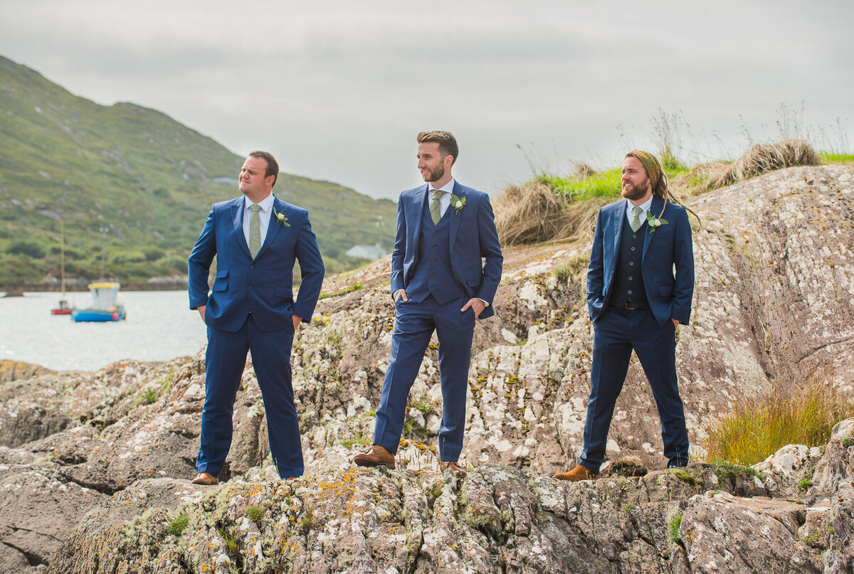 groom and his groomsmen wearing a blue suit and green, tweed tie standing on rocks overlooking Castlecove, Kerry