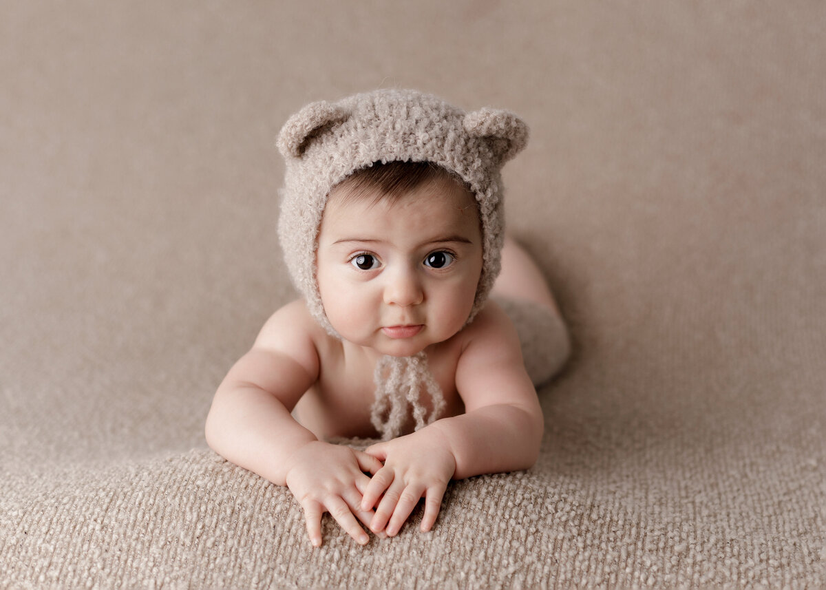 Baby boy laying on his belly for 6-month milestone photoshoot. Baby boy is bare wearing a knit teddy bear bonnet. Baby is propped up on his arms and looking at the camera.