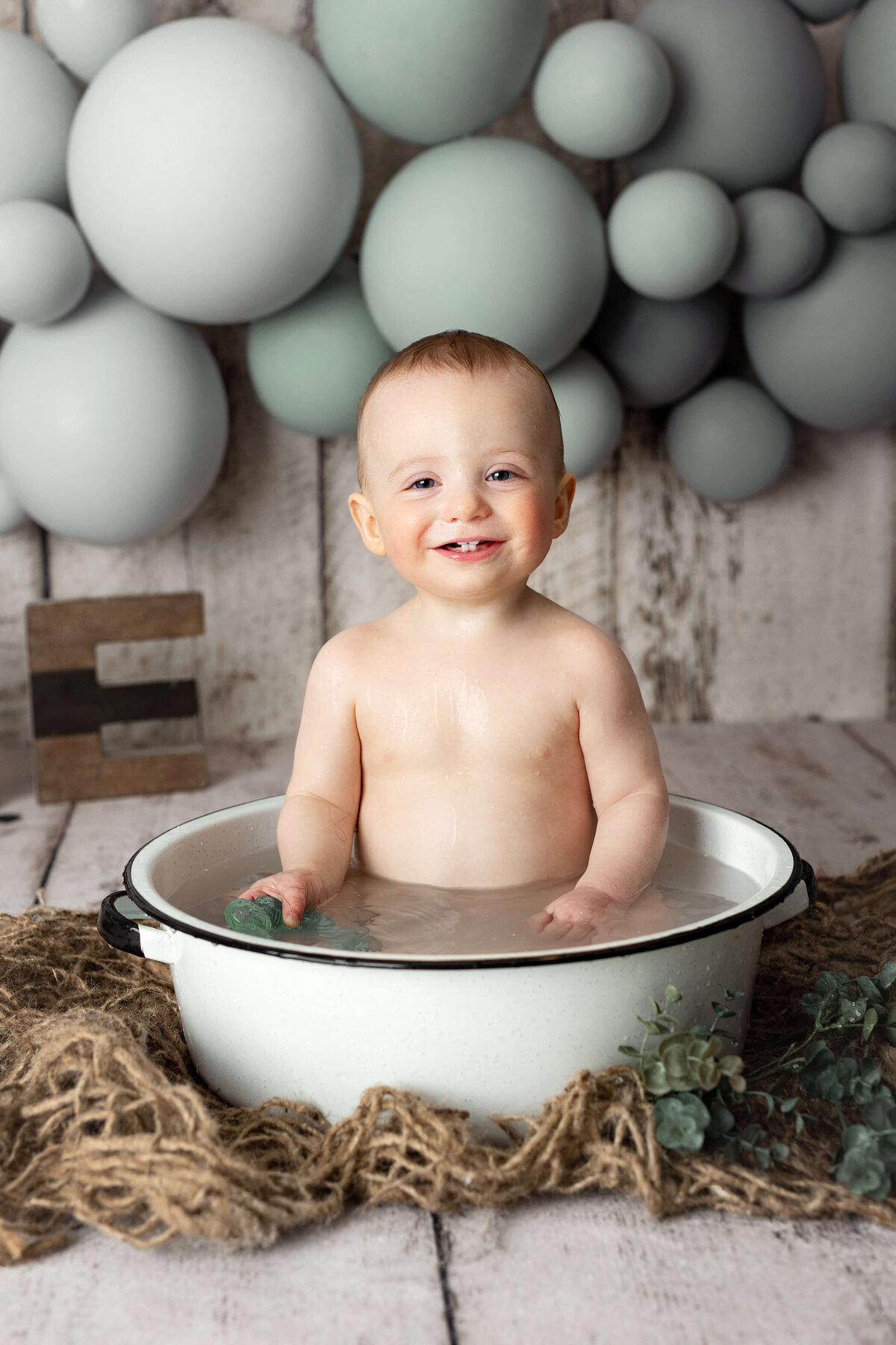 toddler sitting in a bucket filled with water taking a bath with balloons in the background