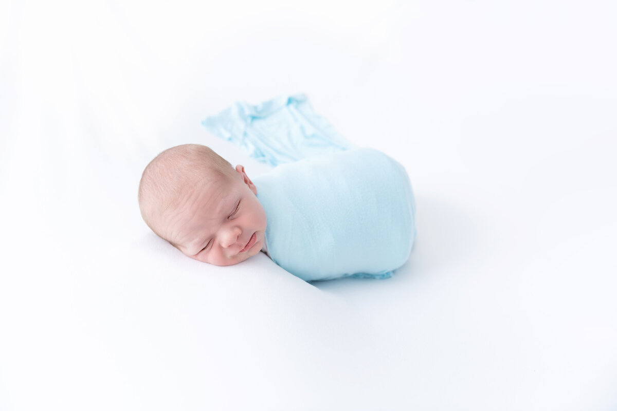 A newborn baby sleeps in a tightly wrapped blue swaddle on a white bed