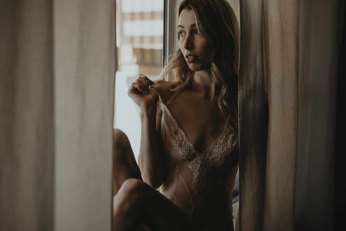 blonde girl sitting on a window cell with her curtains framing her in the middle. She is looking off to the side, out the window and pulling the strap of her body suit away from her body.