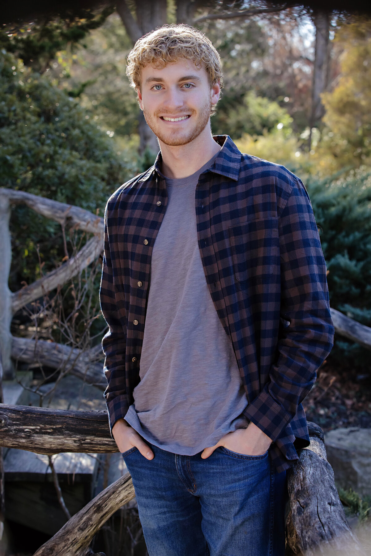 Distinguished autumn portrait of young man in casual button‑down leaning against wooden railing with foliage backdrop at UNCC Botanical Garden Charlotte NC