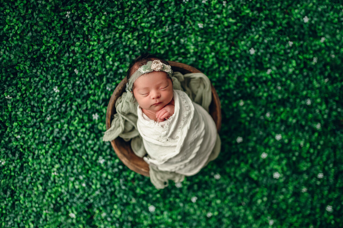 Portrait of a baby girl wrapped in a white, boho-style blanket  and sleeping in a wooden bowl on a boxwood background.