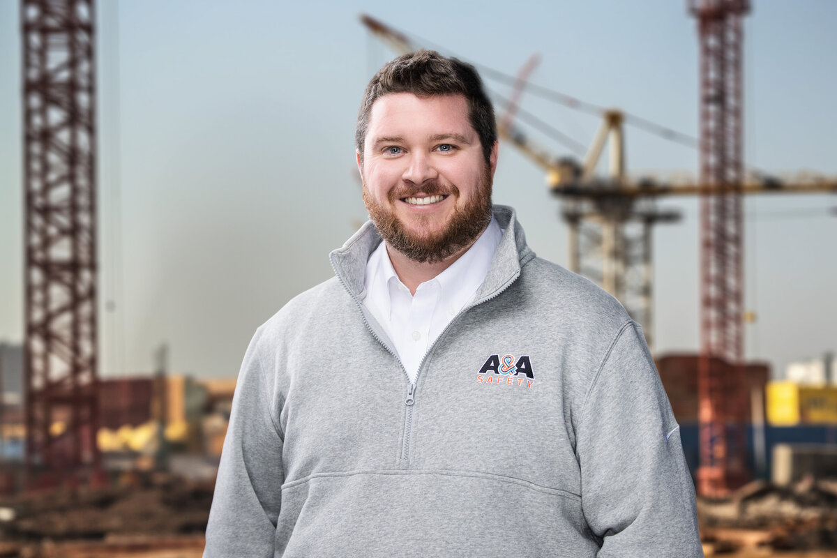 This professional headshot showcases a bearded man with short, neatly styled dark hair. He is dressed in a light gray quarter-zip fleece embroidered with the "A&A Safety" logo over a crisp white collared shirt. The man's friendly smile and confident posture, paired with the construction site backdrop featuring cranes, highlight his professional and approachable demeanor. This headshot, taken in Cincinnati, is perfect for corporate branding and personal marketing needs.