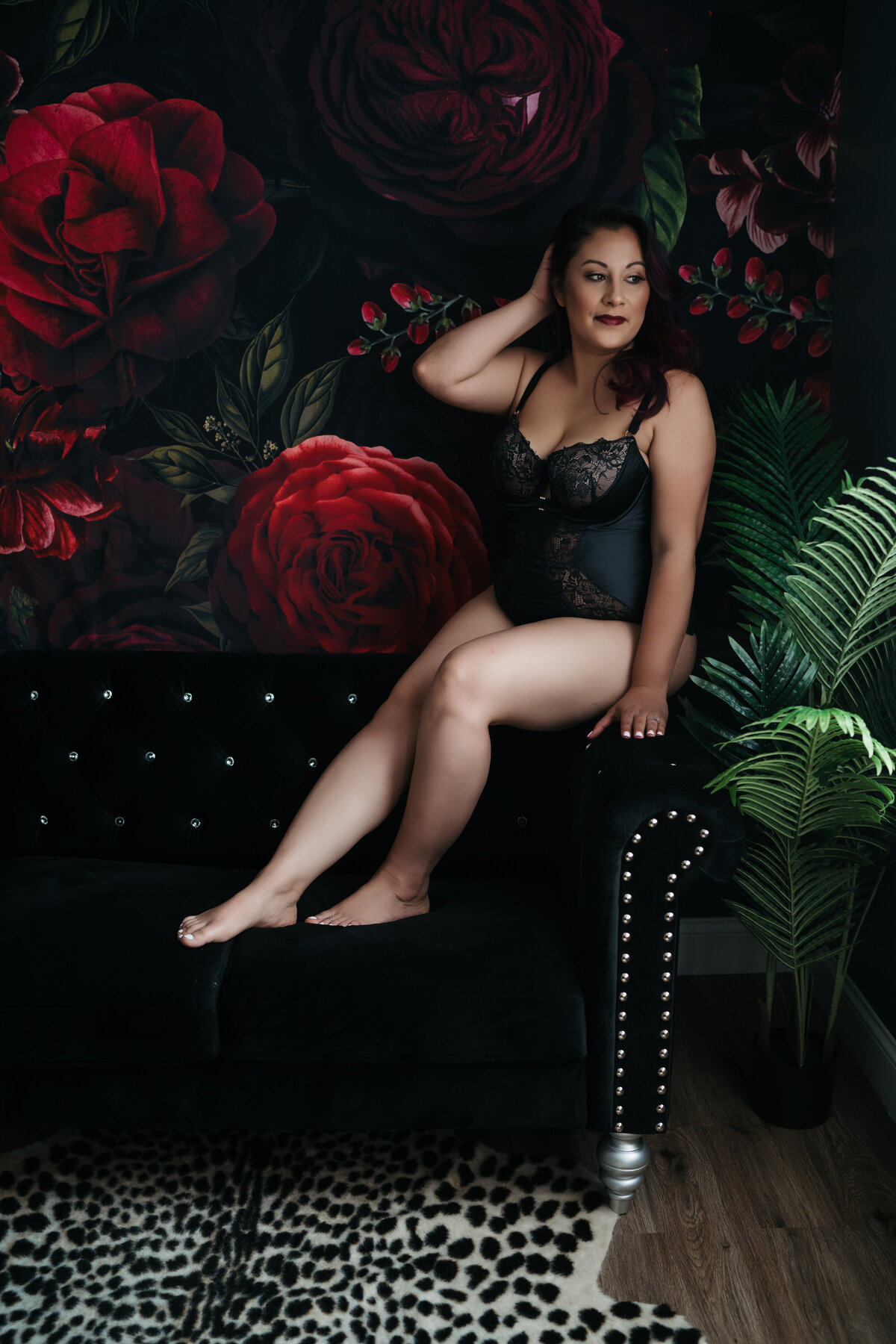 A woman in black lace lingerie sits on the arm of a black chair in front of a large floral tapestry