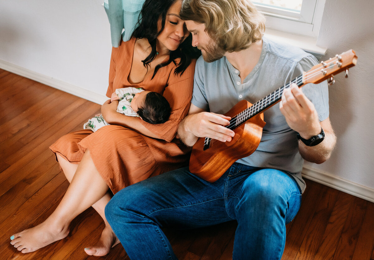 Newborn Photographer, Mom and Dad snuggle and play the guitar while holding baby girl.