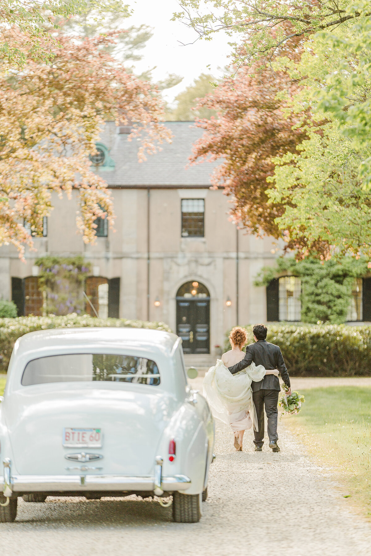 Bride and groom walk with their arms around each other's waist toward a historic building on the North Shore of Massachusetts. In the foreground there is a white vintage Rolls Royce. Captured by best Massachusetts wedding photographer Lia Rose Weddings