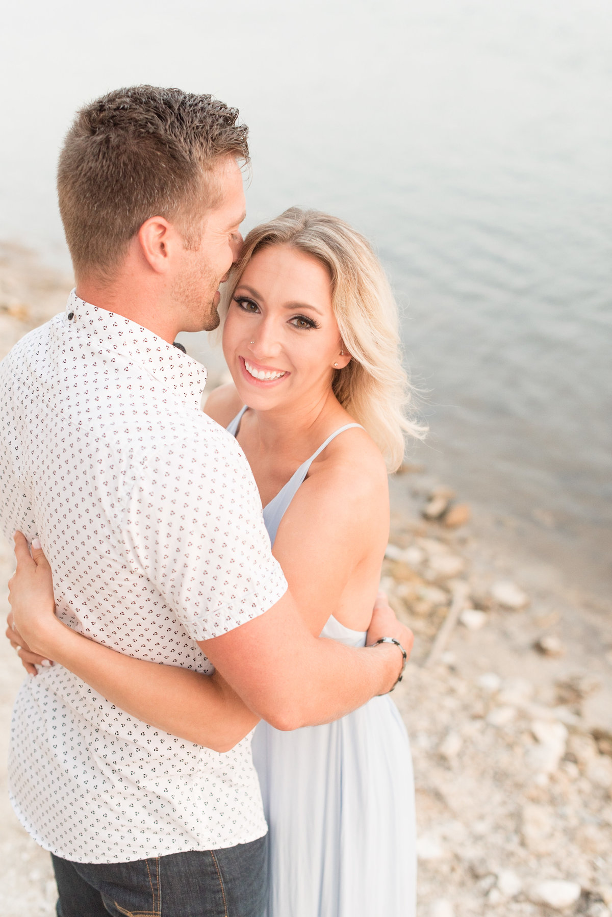 Engaged couple hugging as woman smiles and looks at camera beside edge of pond.