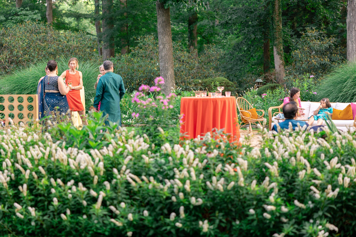 A garden party with several people conversing and seated among lush green plants and flowers. A table with an orange cloth holds drinks. Trees and greenery surround the scene, creating a serene outdoor atmosphere.