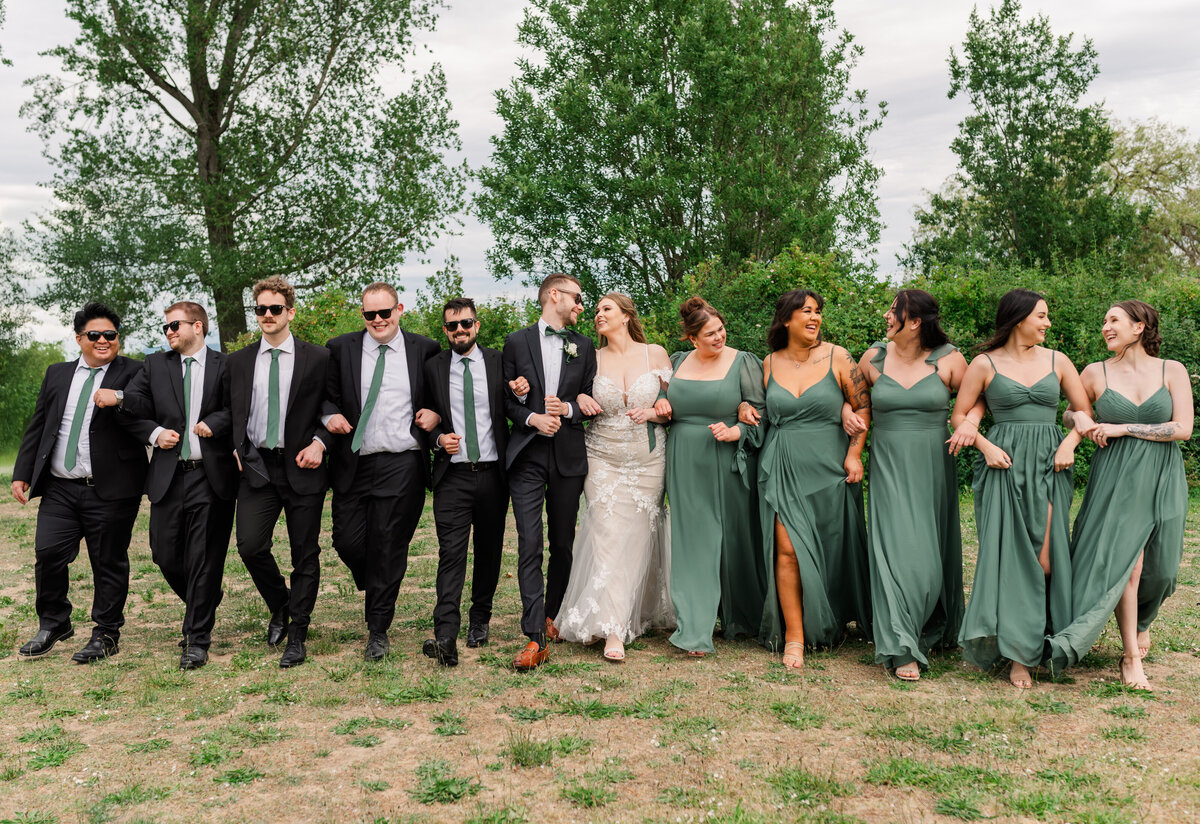 Large wedding group link arms laughing and walking forward, green trees in the back round.