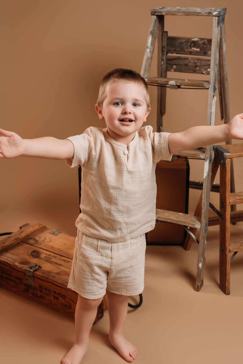 A child in a beige outfit smiles in front of a neutral-toned backdrop featuring  a vintage suitcase and rustic ladder, captured by a photographer in Bemidji, Minnesota.