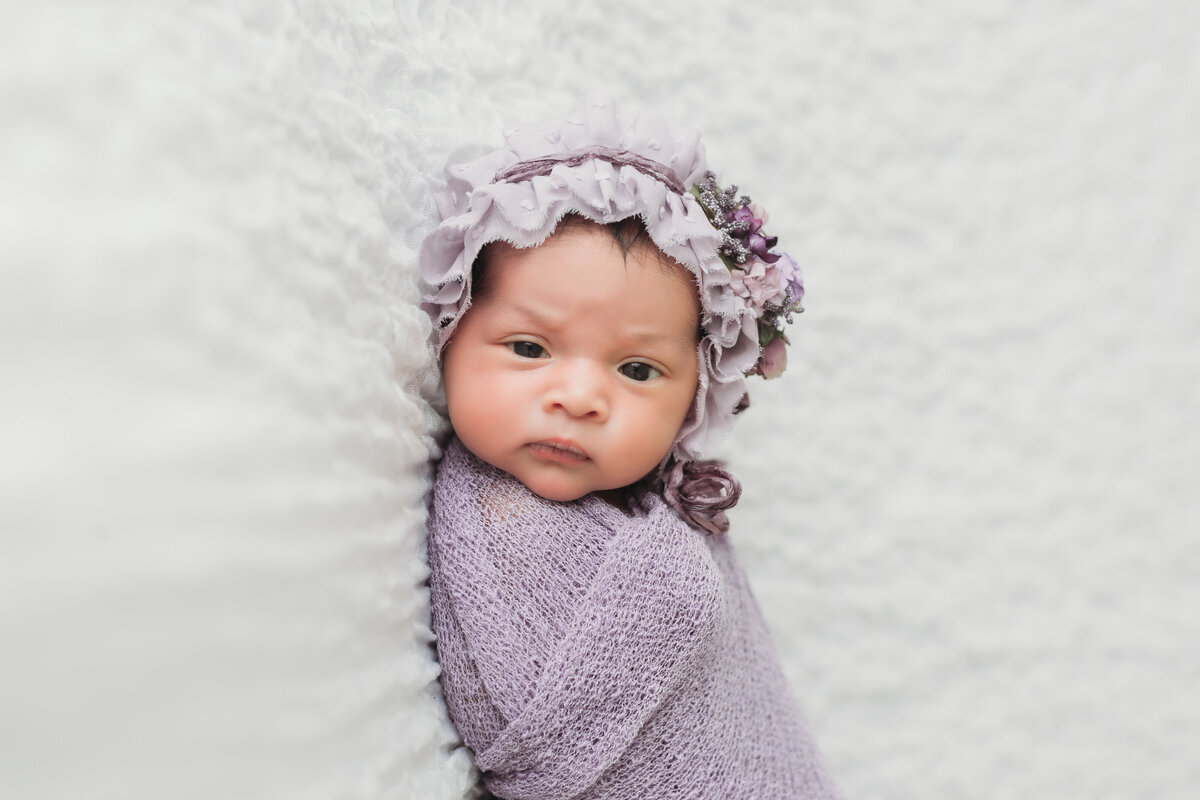 Newborn Photographer, a baby girl sleeps wrapped in purple blankets and wears a cap with flowers on it
