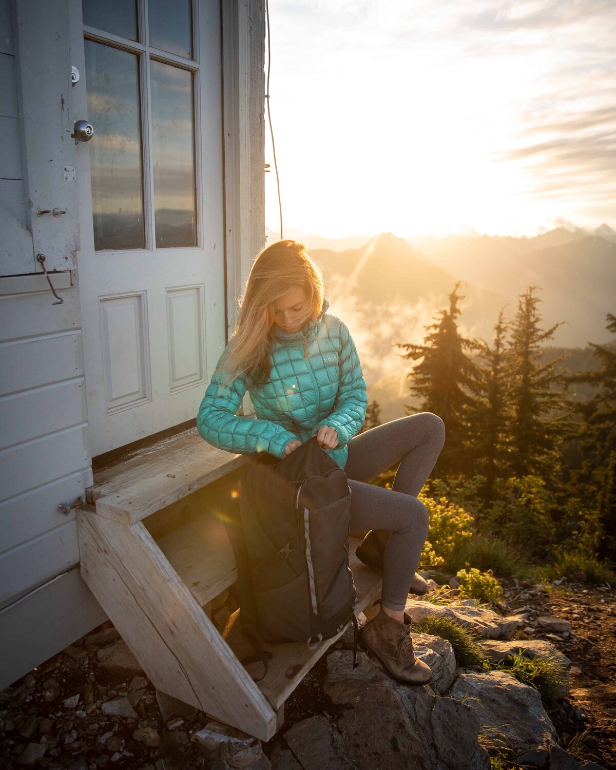 Jess of Jess Wandering in a blue Backcountry jacket sitting on the steps of a house with the sun rising in the background