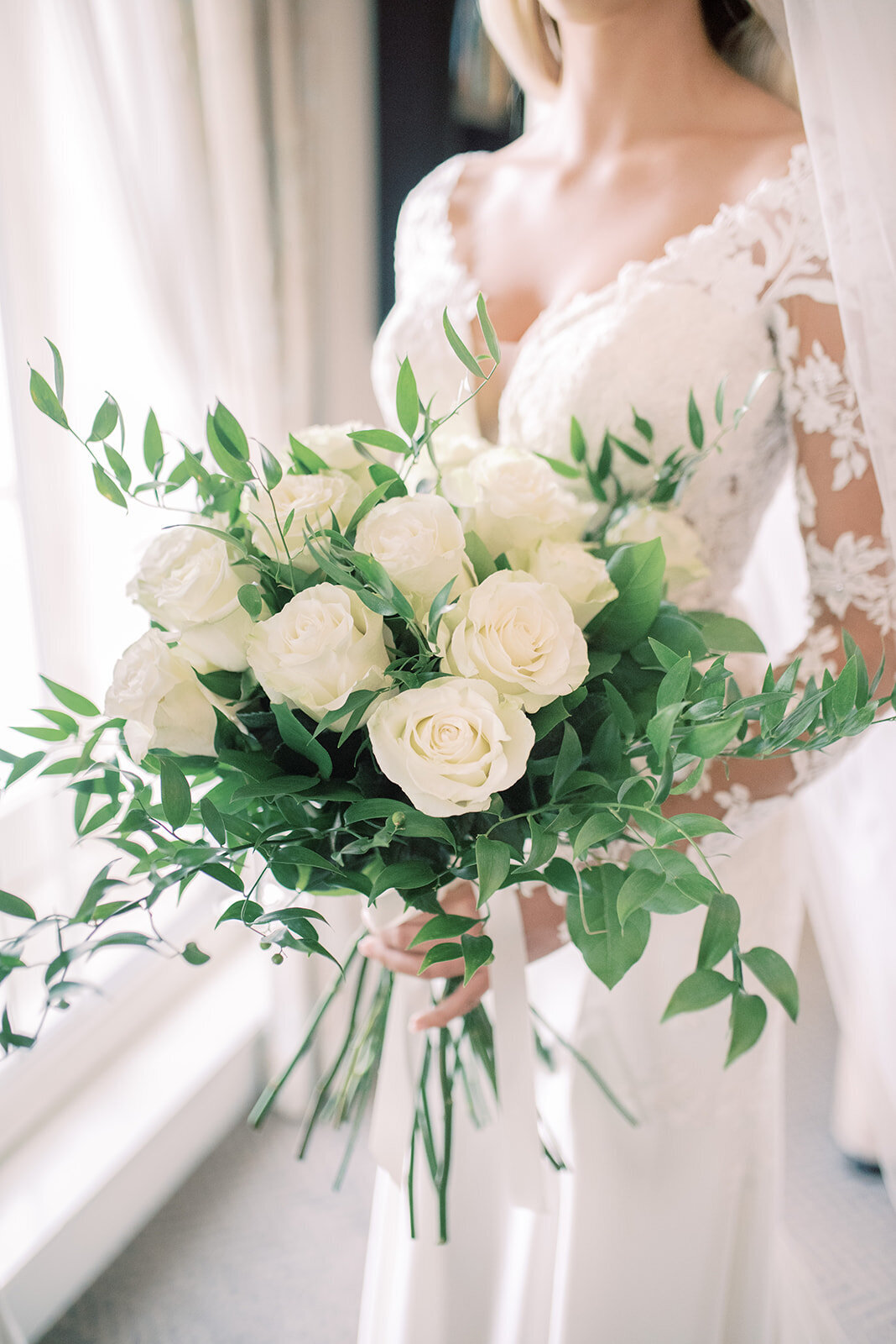 Bride holding her bouquet stood by the window, the bouquet is white long stem roses and the bride is wearing a white lace gown. The image is focused on the bouqet