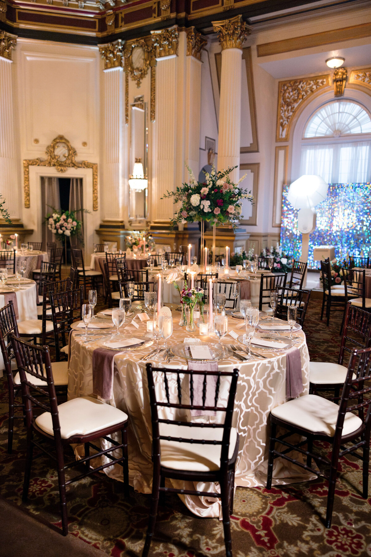 A beautifully decorated wedding reception with round tables covered in elegant white and silver tablecloths, adorned with candles and floral centerpieces. The room features high ceilings, ornate gold detailing, and chairs arranged around the tables.