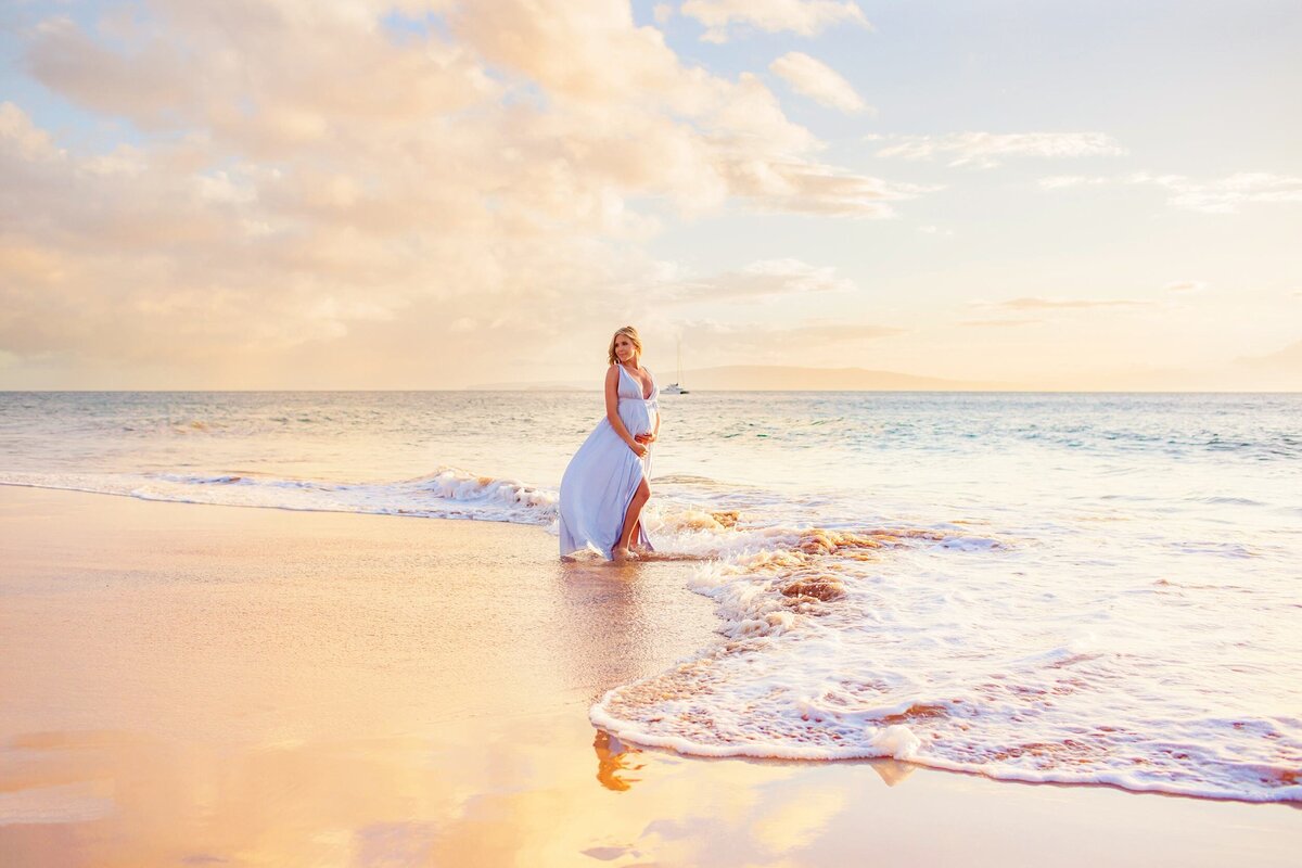 Woman in blue dress poses at sunset during her maternity session in Wailea as waves lap on the shoreline