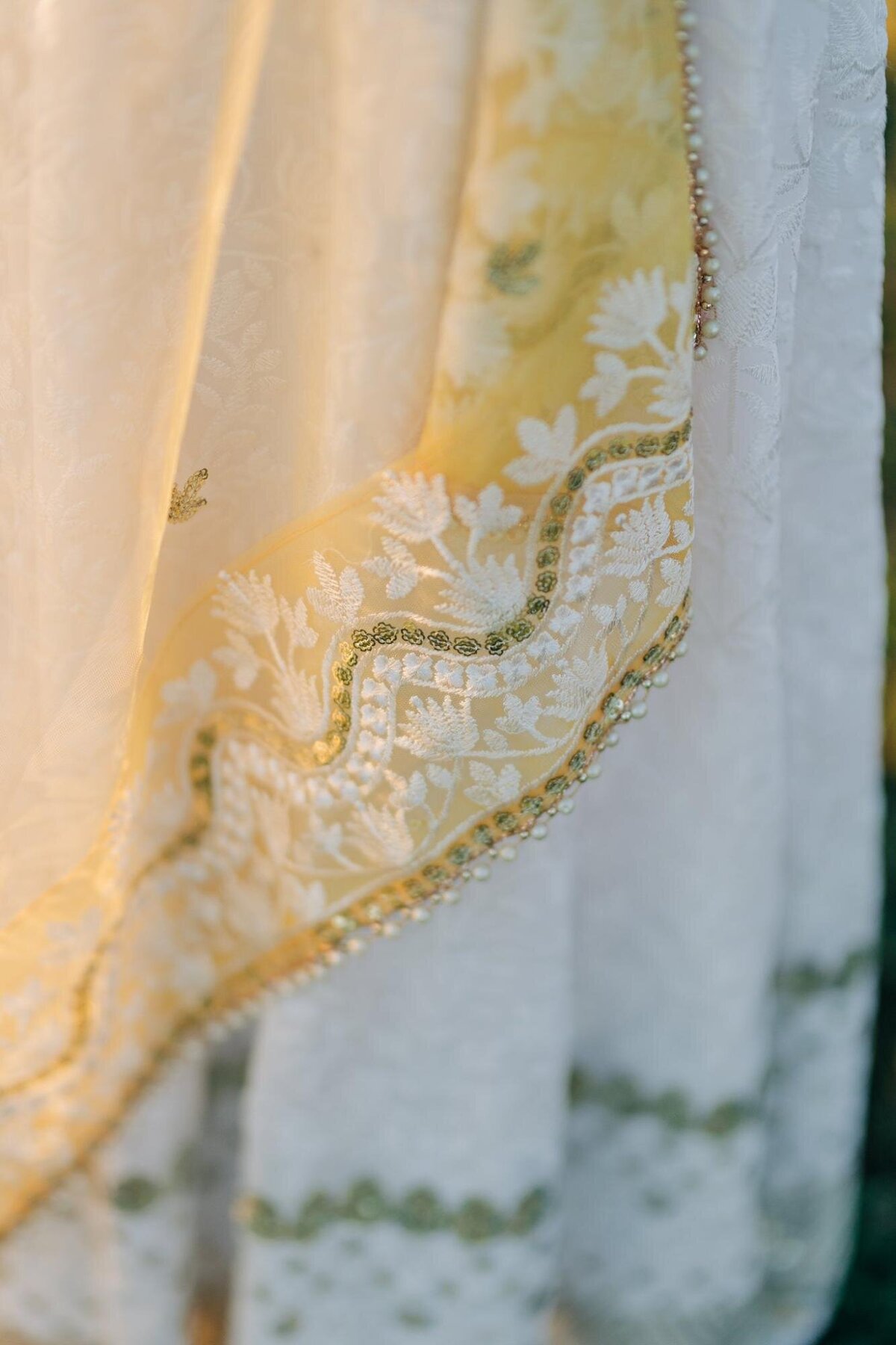 Close-up of a delicate white and gold embroidered fabric with intricate beadwork, highlighted by soft sunlight.