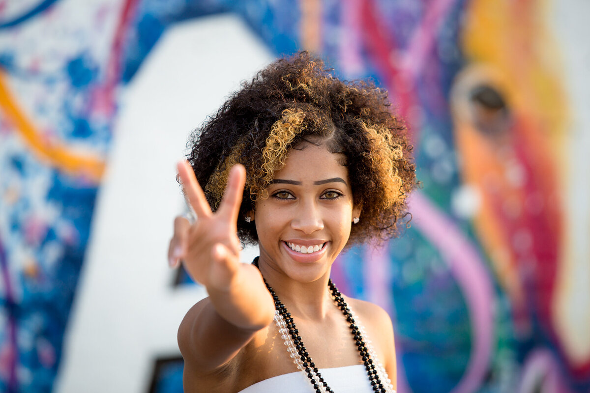 senior girl standing against  graffiti wall giving peace sign