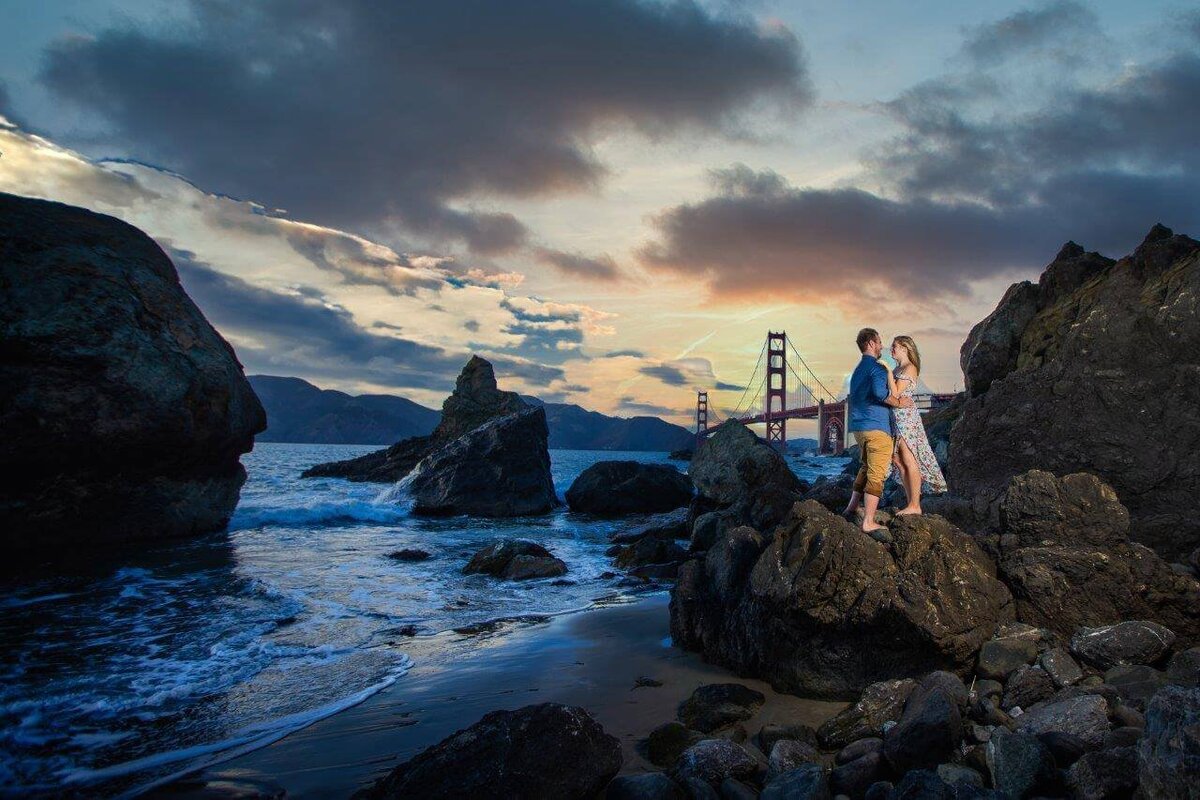 Philippe studio pro, a wedding photographer from sacramento captures an engaged couple in front of the golden gate bridge standing on rocks with the ocean water surrounding them.  They face each other and look at each other lovingly.