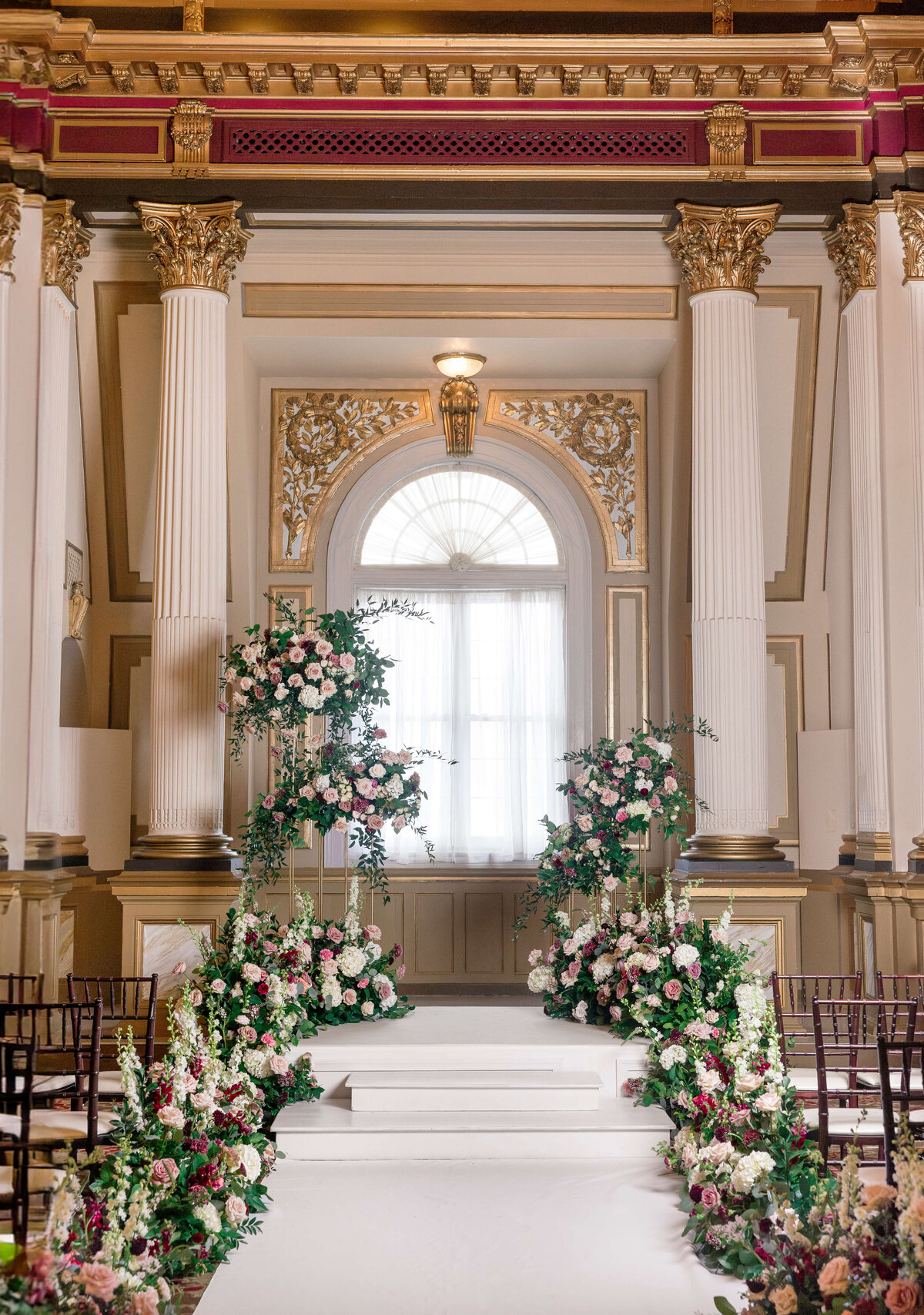 An elegant indoor wedding altar featuring tall white columns and high ceilings. Floral arrangements with pink and white flowers adorn the steps leading to a large window with golden accents. Rows of chairs line the aisle.