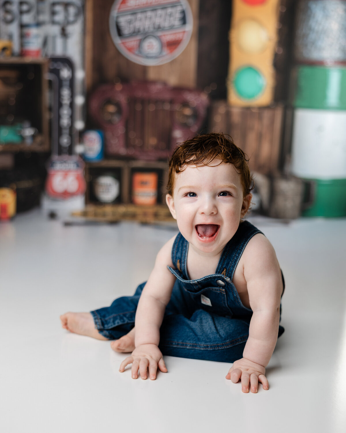 Happy baby boy in denim overalls smiling big while sitting in front of a vintage garage-themed birthday backdrop.
Photograph by Yucaipa cake smash photographer Kaitlyn Dawn Photography.