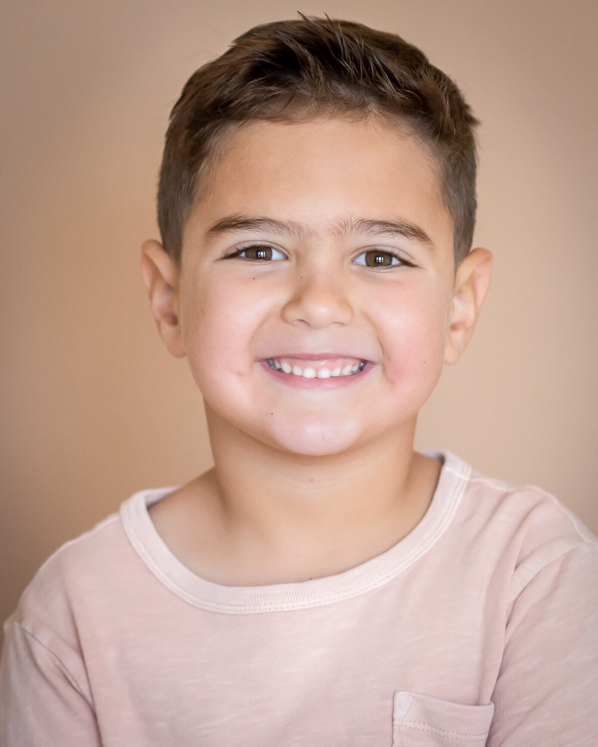 close up portrait of smiley uoung boy in a muted pink tee shirt with a tan background