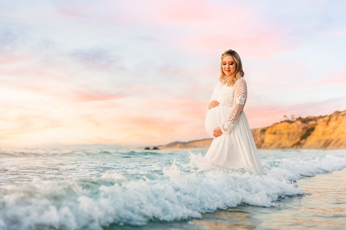 Woman holding pregnant belly and looking down while waves splash around her on the beach in La Jolla with a view of Blacks Beach behind her