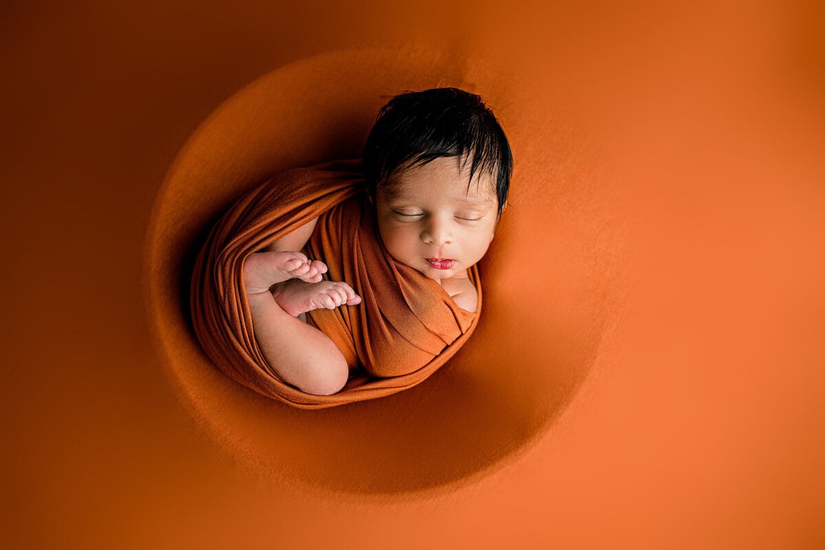 infant baby is wrapped in a burnt orange wrap and laying on a matching drop in a charlotte studio