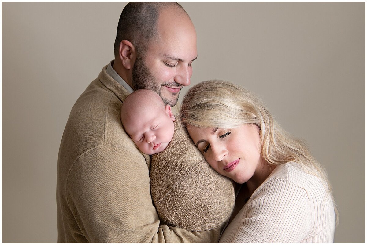 Close up studio image of dad holding his baby boy and mom is resting her head on the baby with her eyes closed.