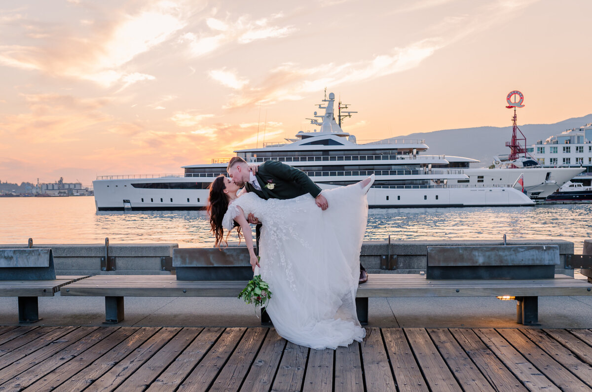Bride dipped down one leg out and groom holding bride up while kissing. Vancouver shipyard with big boats in the back round.