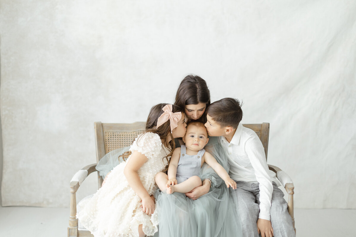 A photo of a mother and her 2 young children sitting on a bench in a photography studio as they all lean in to kiss the baby.