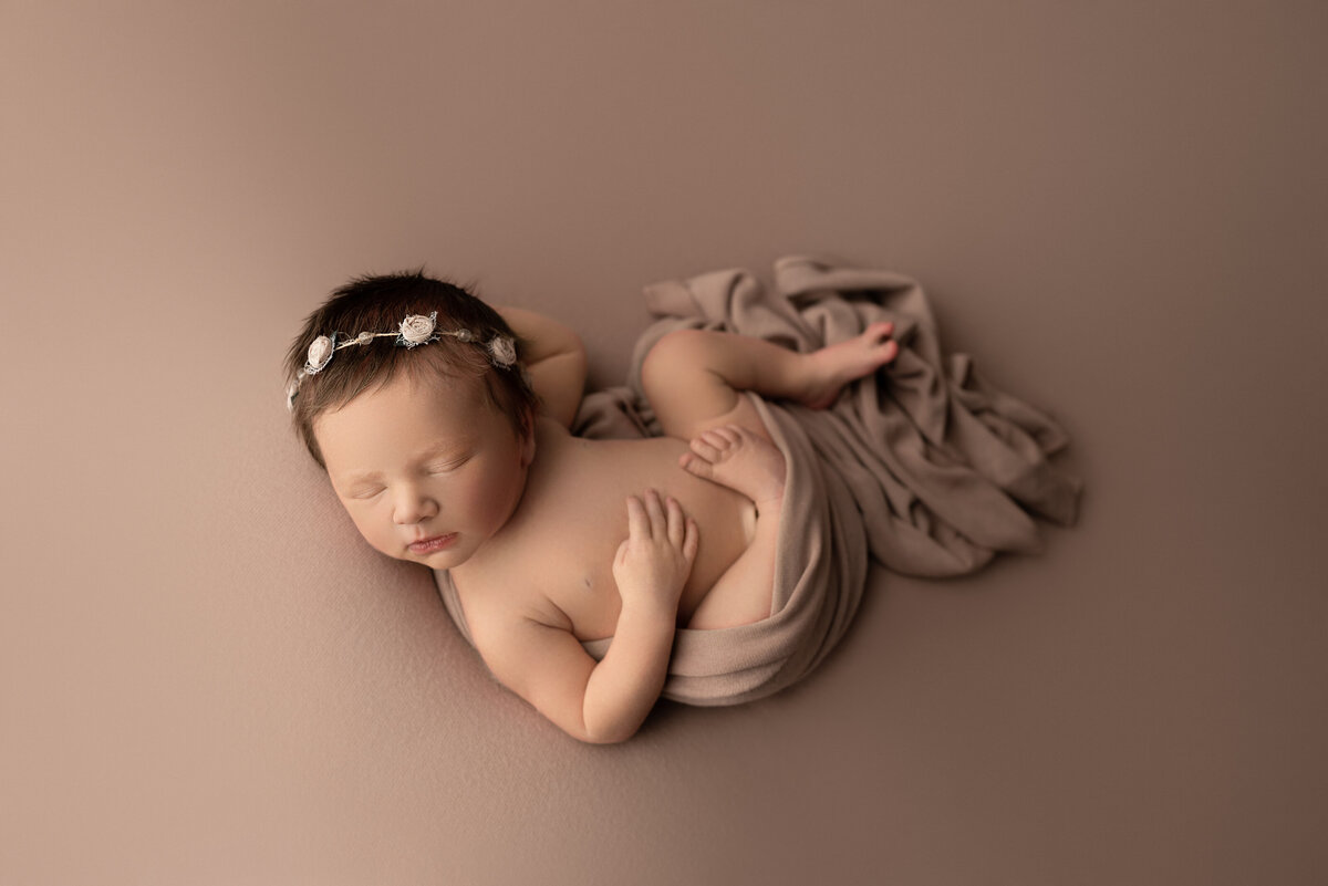 Philadelphia's best newborn photographer, Katie Marshall captures a fine art newborn image. Baby girl is sleeping on her back with a fabric swaddle wrapped underneath her. Her legs are folded up. One of baby's hands are resting on her belly, the other underneath her head. Baby is wearing a headband with delicate flowers. The image is captured at an angel with baby's head turned toward the camera.