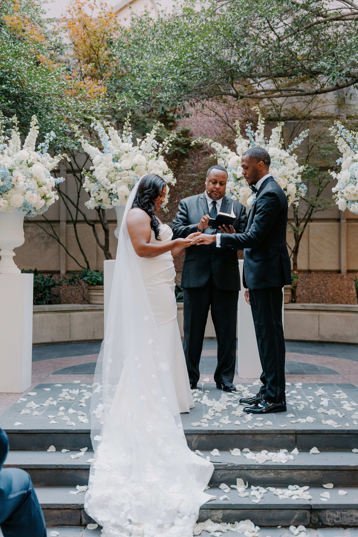 Bride and groom exchange rings at a wedding ceremony, with an officiant standing between them. They are surrounded by white floral arrangements, exuding joy.