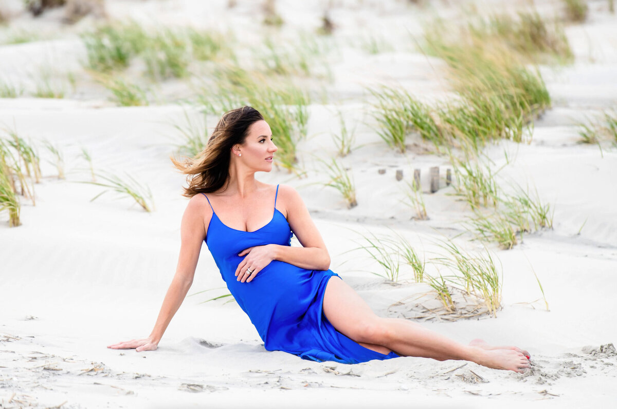 Woman posed for a maternity photo, wearing a satin blue dress sitting on the beach in Stone, Harbor New Jersey