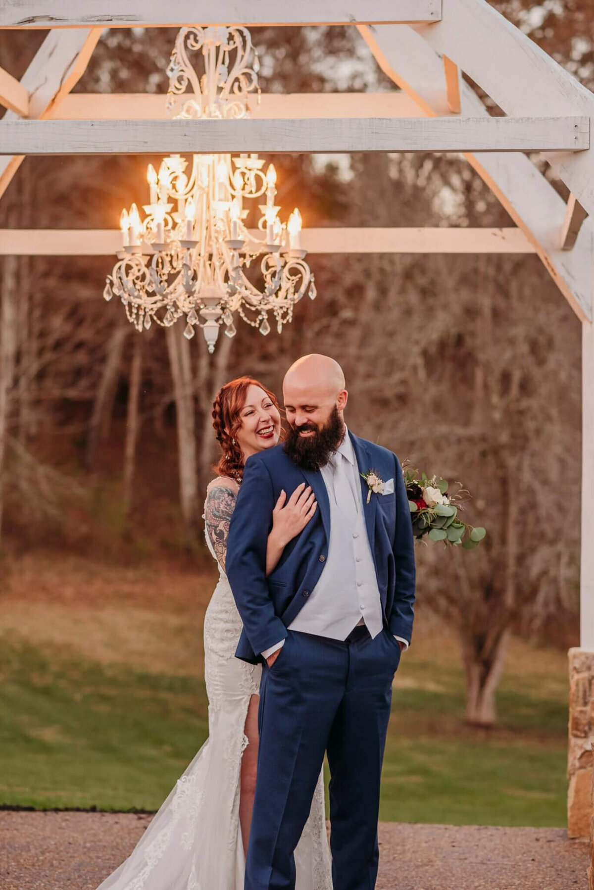 photo of a bride hugging a grooms back while they laugh and stand under a chandelier