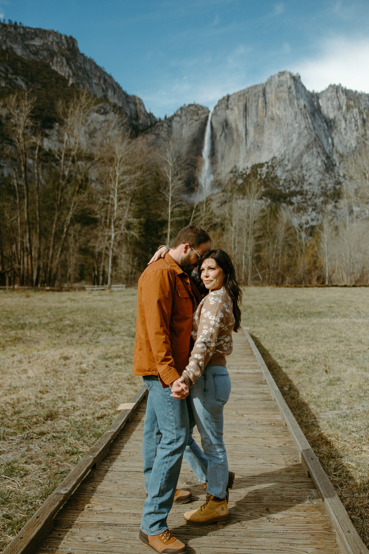 A couple on a pathway holding hands and facing eachother in fall with a beautiful waterfall and rock formations in the back