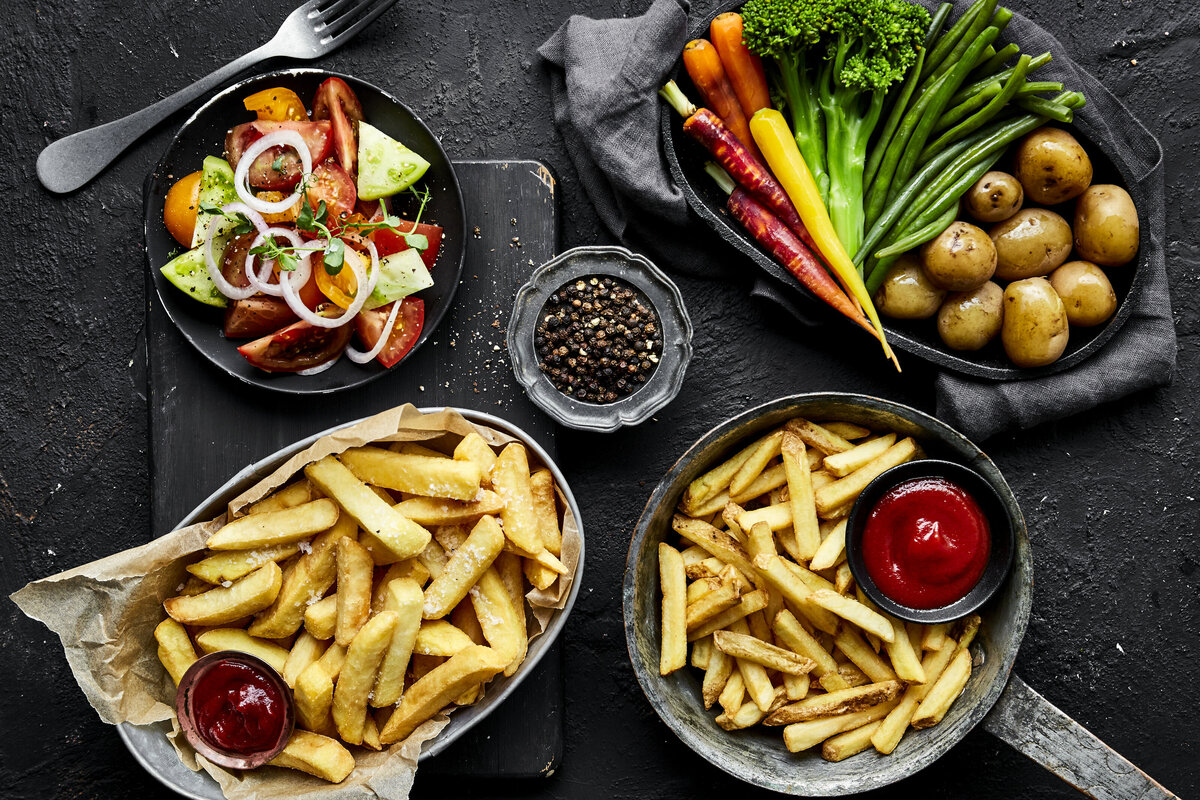 Two plates of fries with plates of vegetables on the table next to them.
