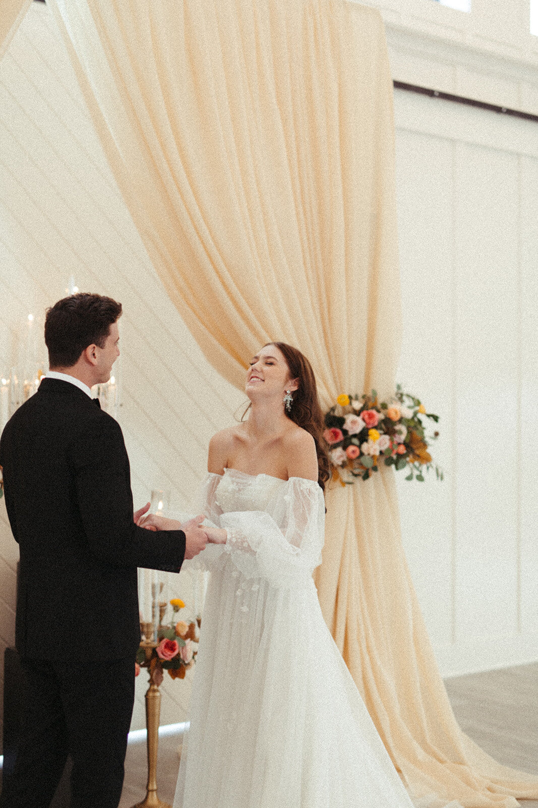 A groom wearing a black tuxedo holds bride wearing a white wedding gown by the hands in front of peach curtains with flowers.