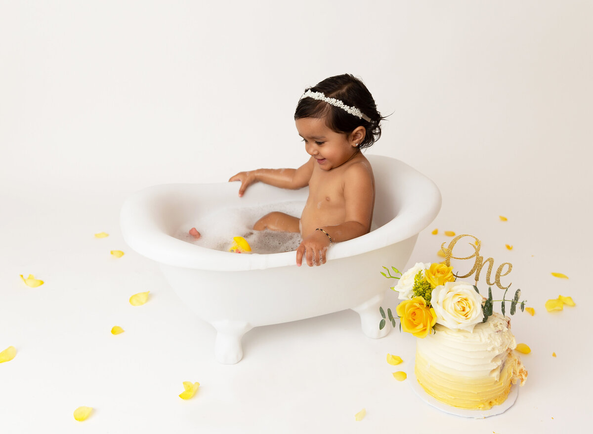 Baby girl sits in a bathtub to clean up from cake smash photoshoot. In front of the tup there is the smashed yellow ombre cake.