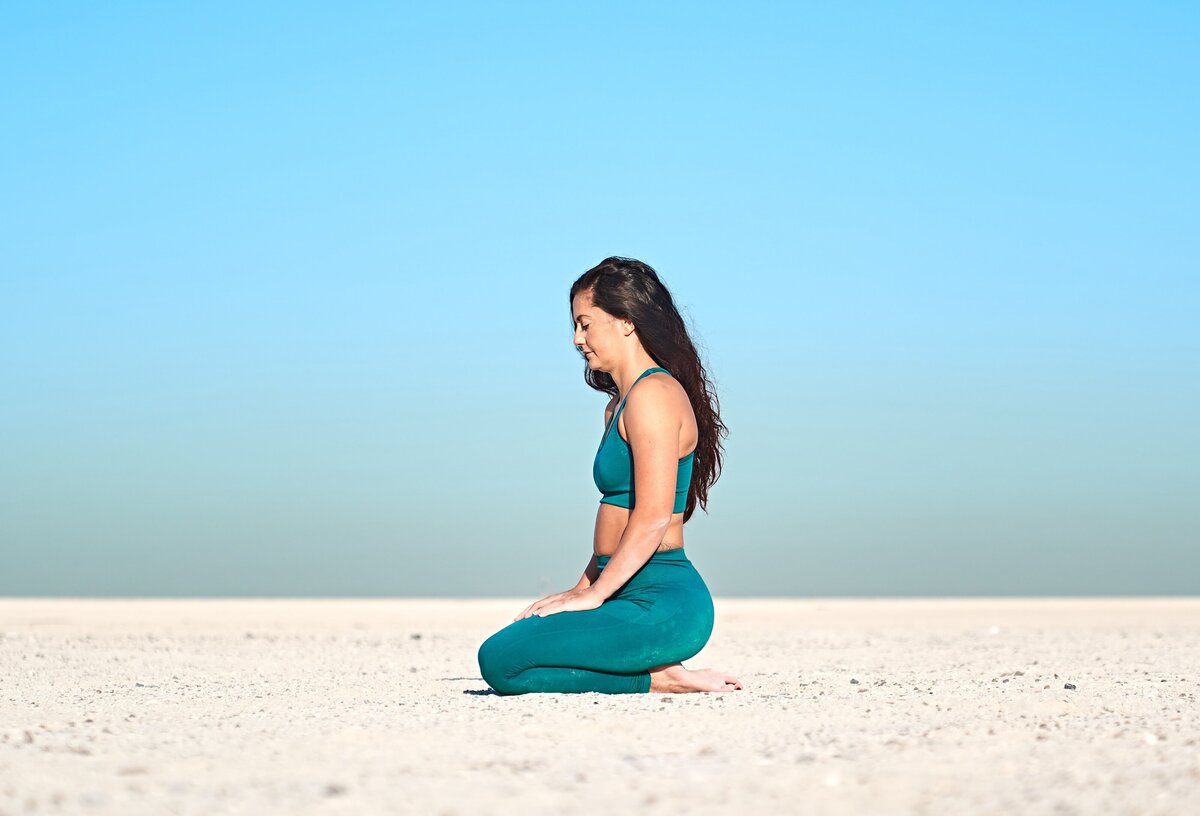 Yoga instructor Sarah sits in Vajrasana, or Diamond Pose, on a stark desert expanse, under a clear sky, encapsulating serenity and groundedness in her surroundings.