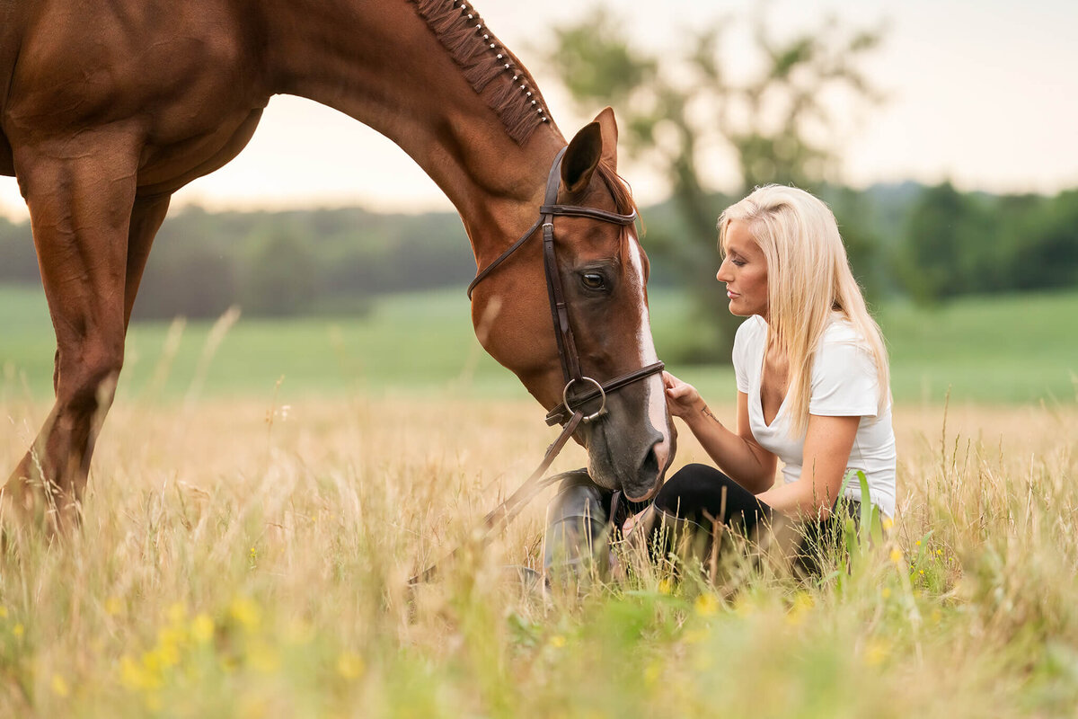 blonde equestrian wearing a white shirt and black shirt sitting in a field with her chestnut quarter horse nuzzling her lap sweetly. behind them is pastures with green grass, trees, and golden hour sun.