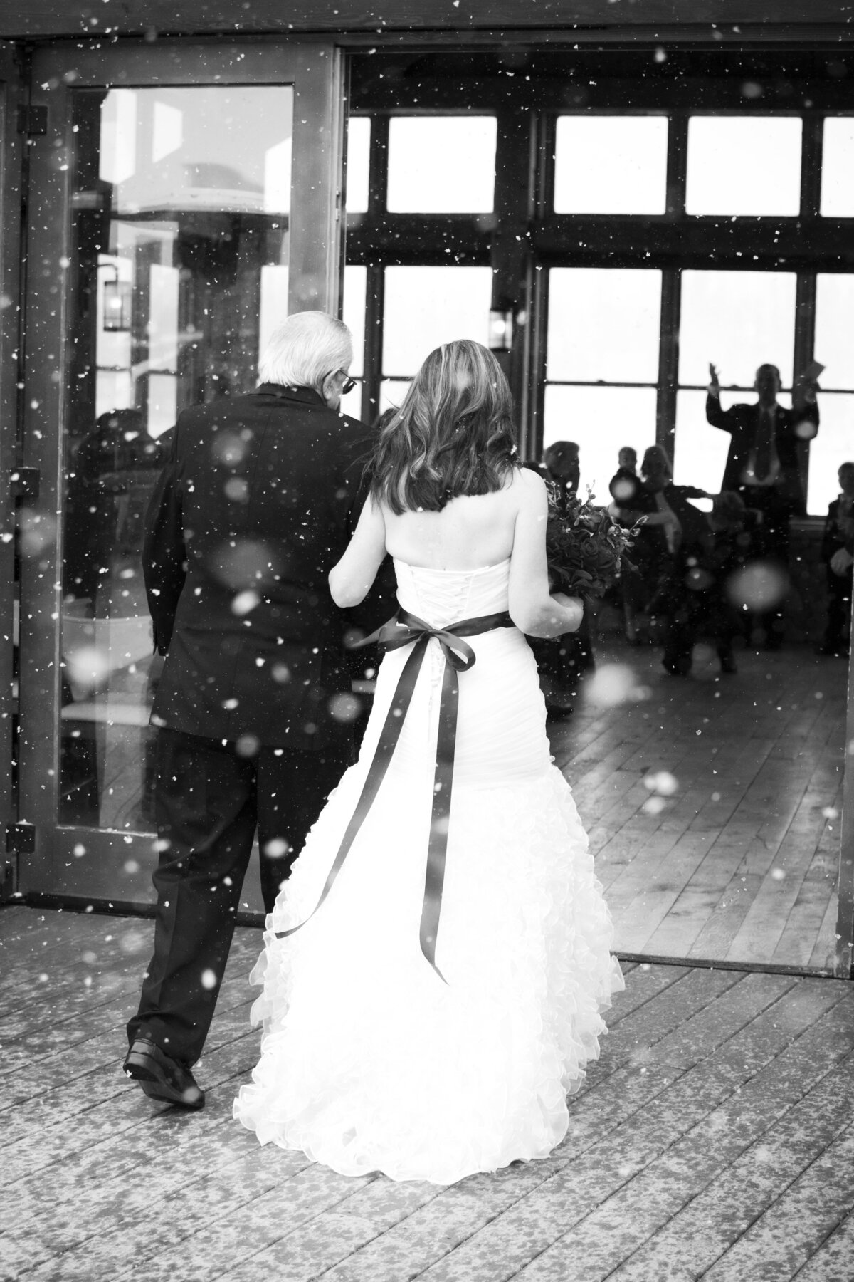 A shot of the bride and her father from behind as they walk up the aisle with snow falling in the foreground.
