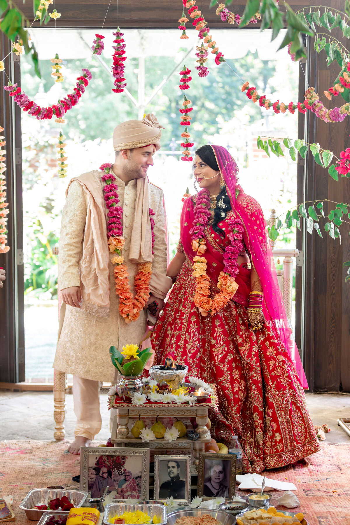A couple in traditional Indian wedding attire stands together, smiling. The groom wears a cream sherwani and turban, while the bride is in a red lehenga with pink dupatta. Theyre adorned with floral garlands, surrounded by colorful decorations and offerings.