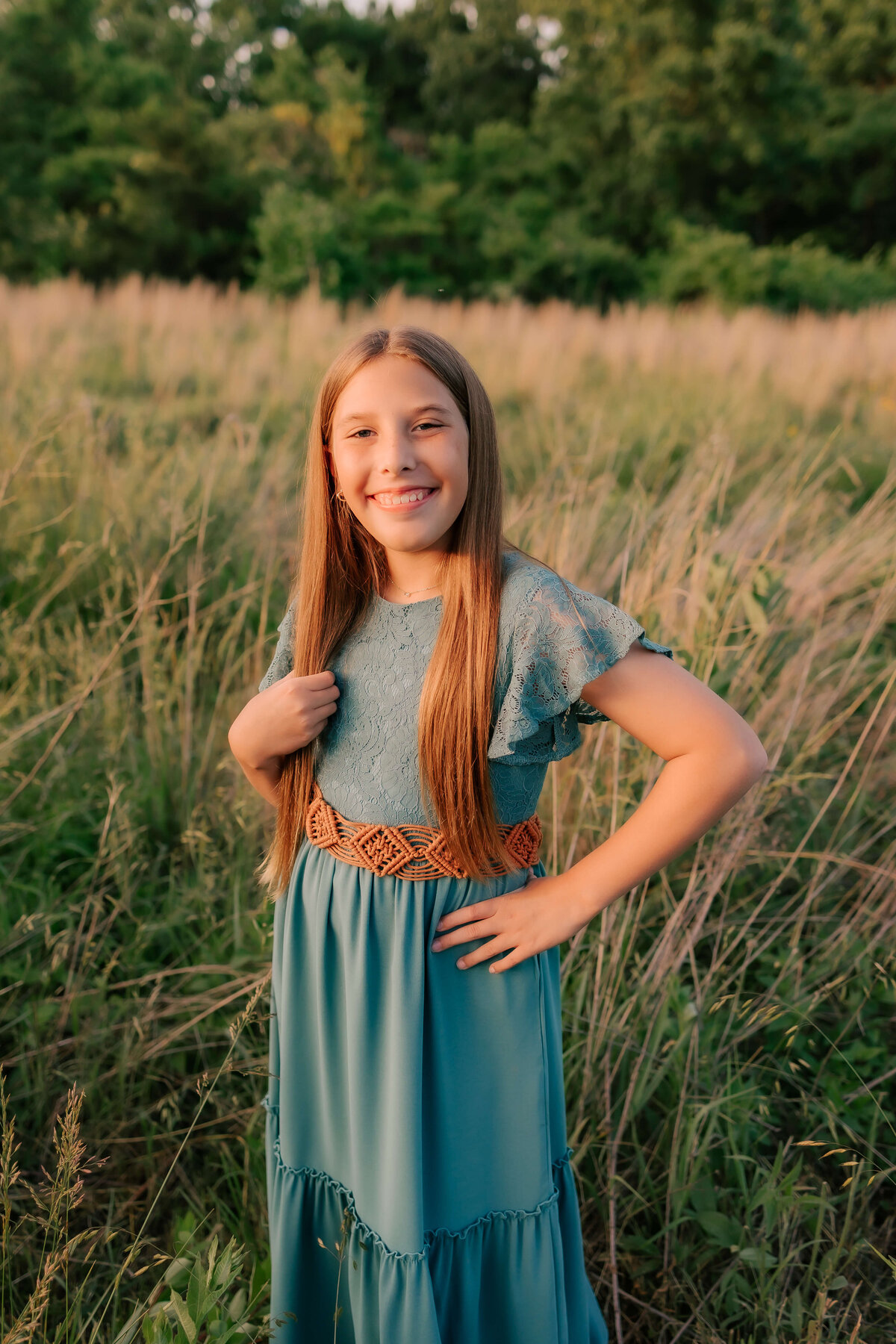 A young girl with long blonde hair is standing in the tall grassy field smiling at Foppiano Photograhy while she captures the beauty of her youth at sunset.