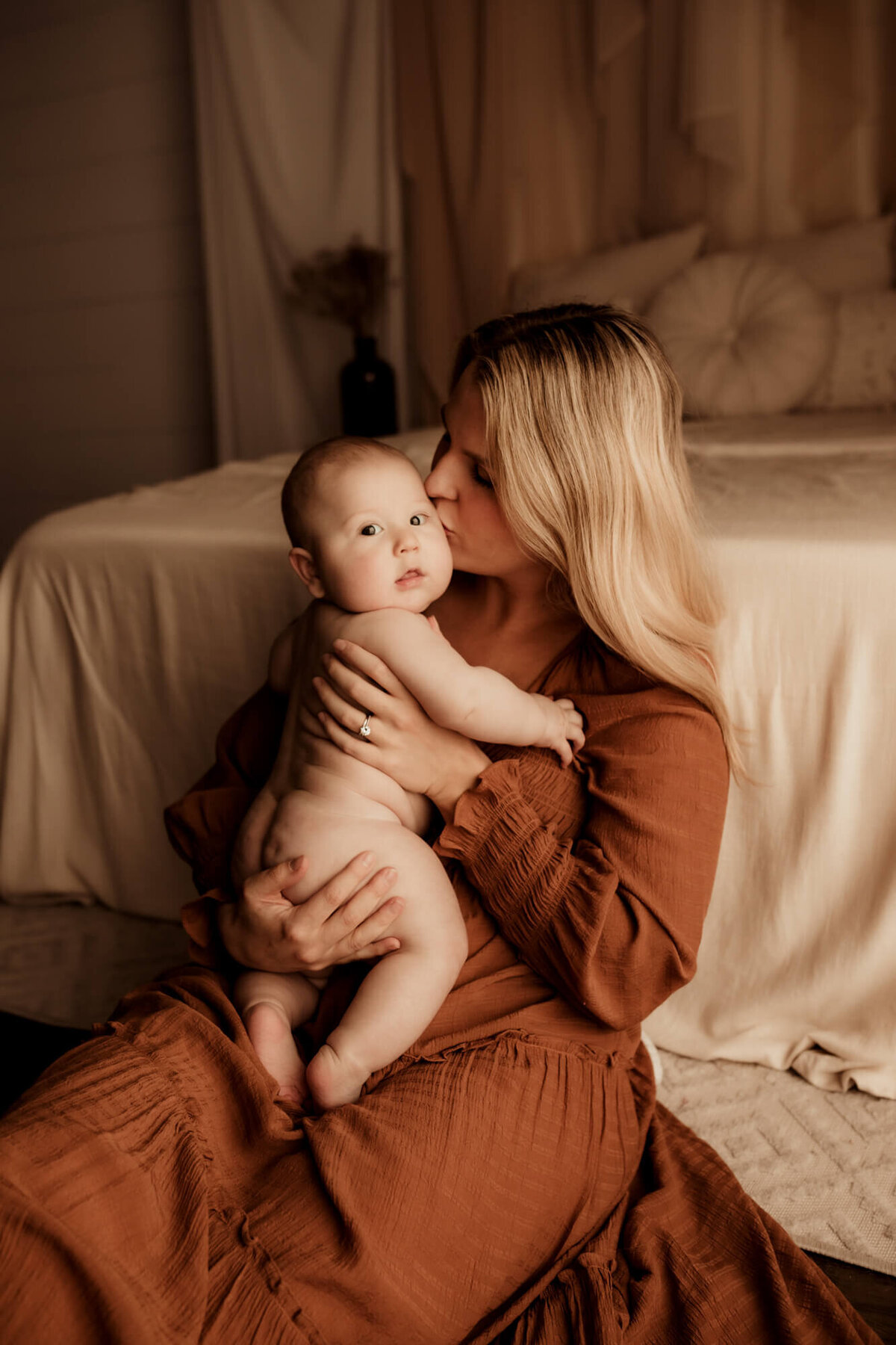 Mother wearing a rust colored dress kisses her baby boy on the cheek.