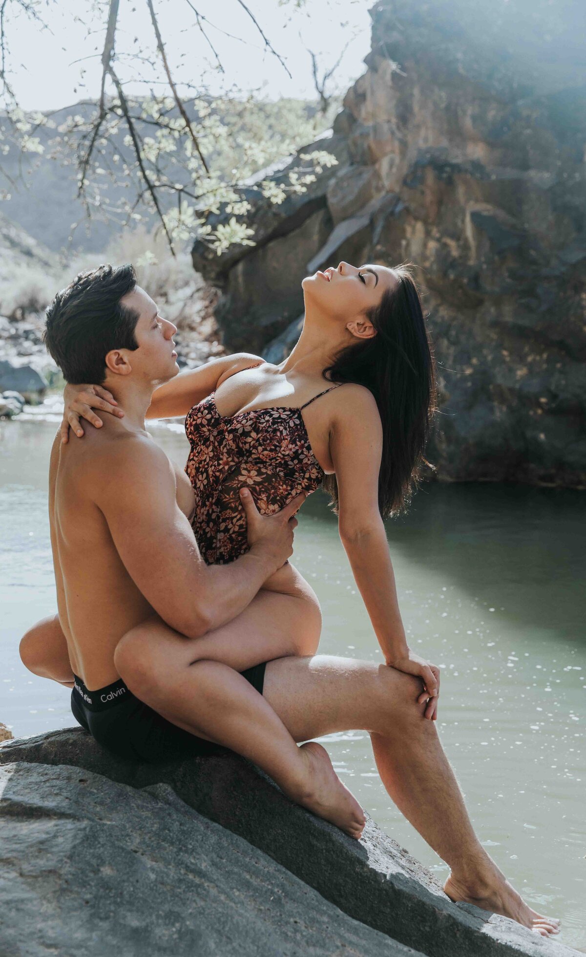 A couple posed on a rock. She has her legs wrapped around him and she is throwing her head back while he holds her.