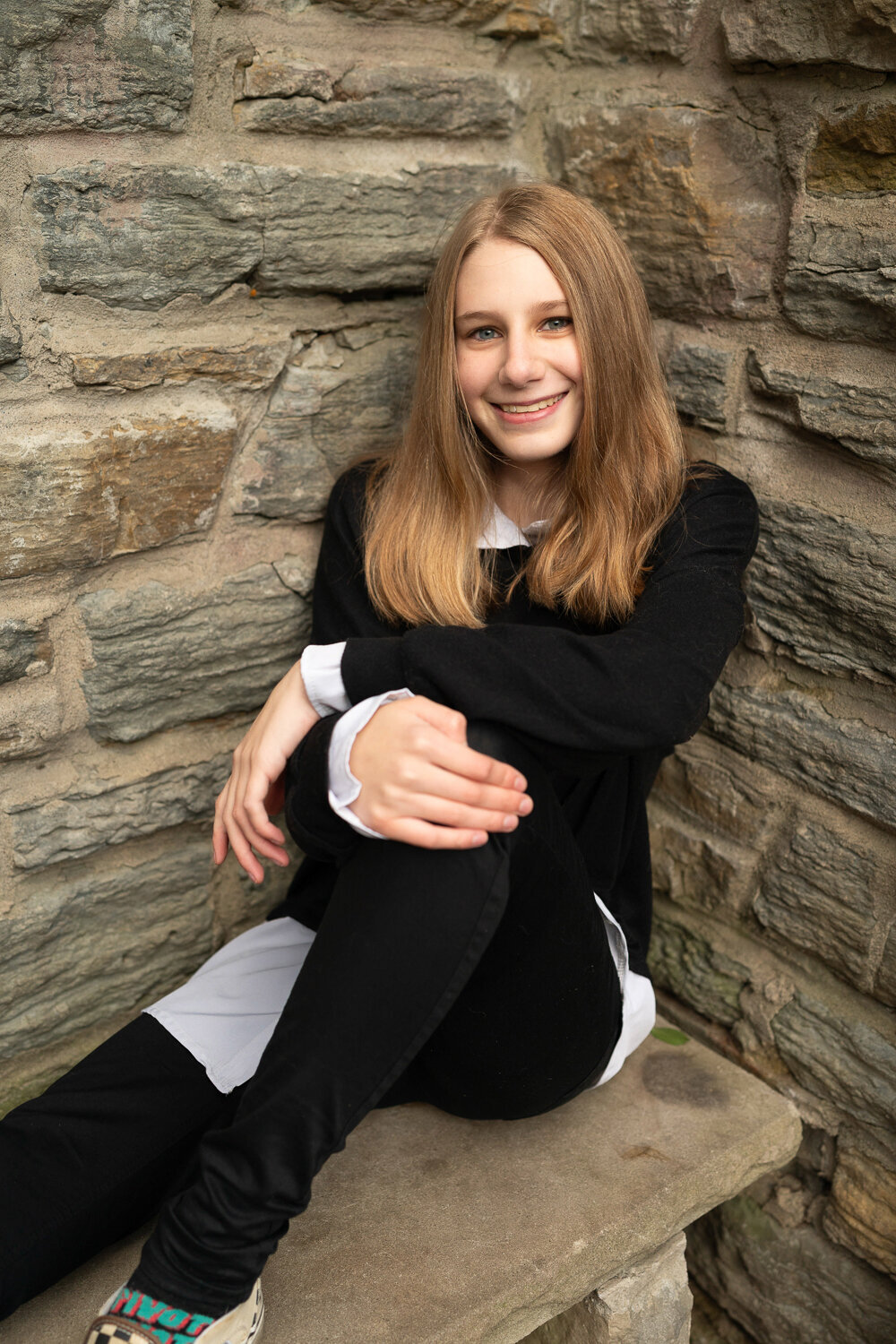 Senior girl in black sweater sits on bench with knee pulled up to chest.