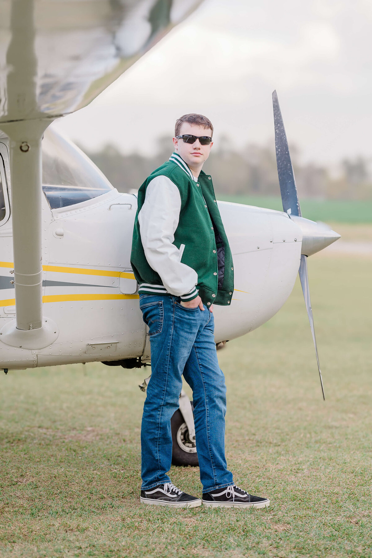 A person in sunglasses and a green varsity jacket leans against the nose of a small white airplane with a propeller. They are standing on grass, with overcast skies in the background.