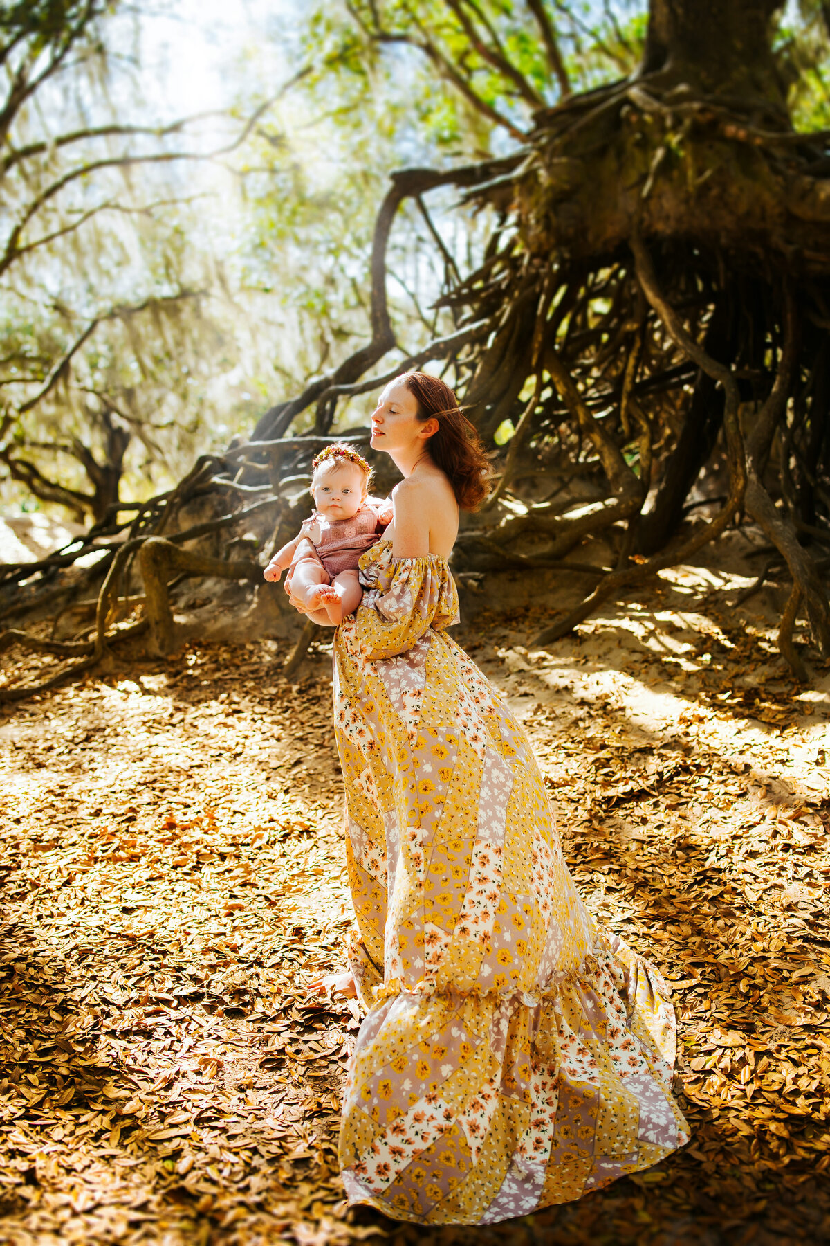 a mother in a floral boho dress with the wind blowing her hair while she's holding her baby during a 1 year photo shoot